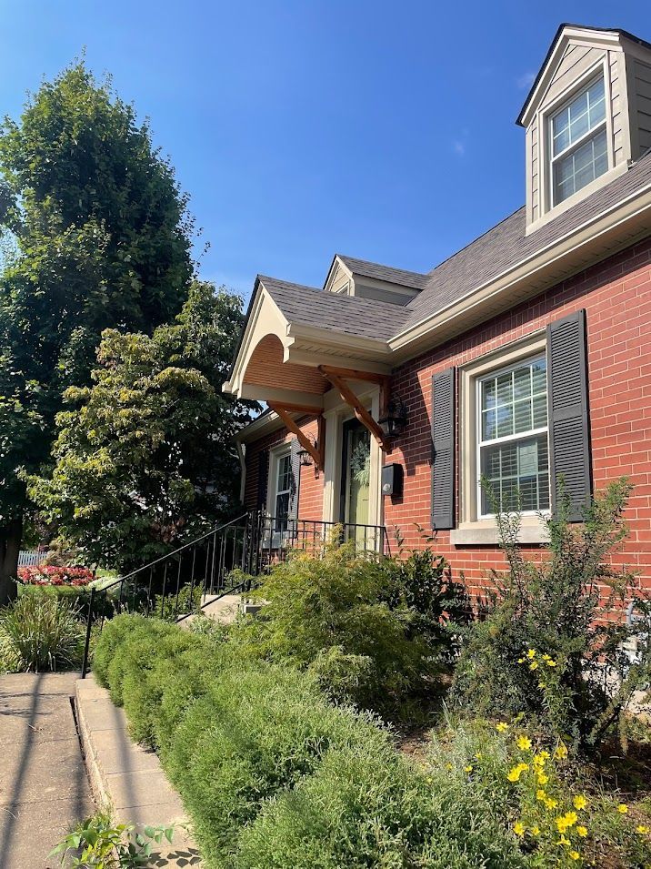 Red brick house with dark shutters, wooden awning, and lush greenery on a sunny day.