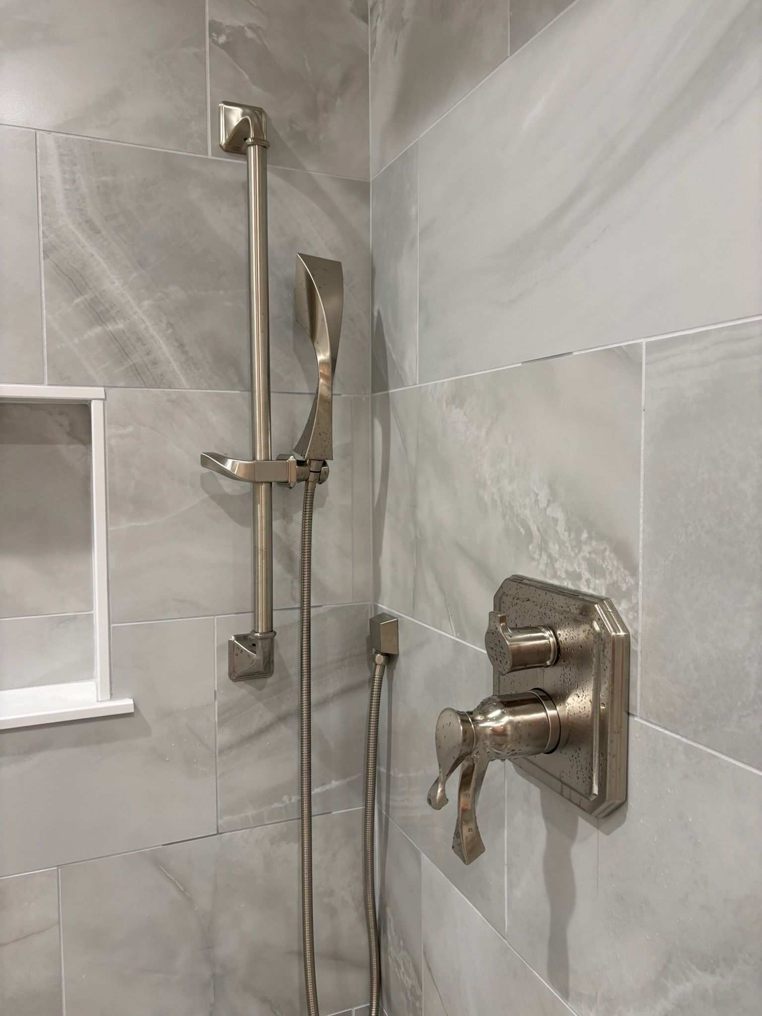 Shower with gray tile, brushed nickel fixtures, adjustable hand shower, and a built-in shelf.