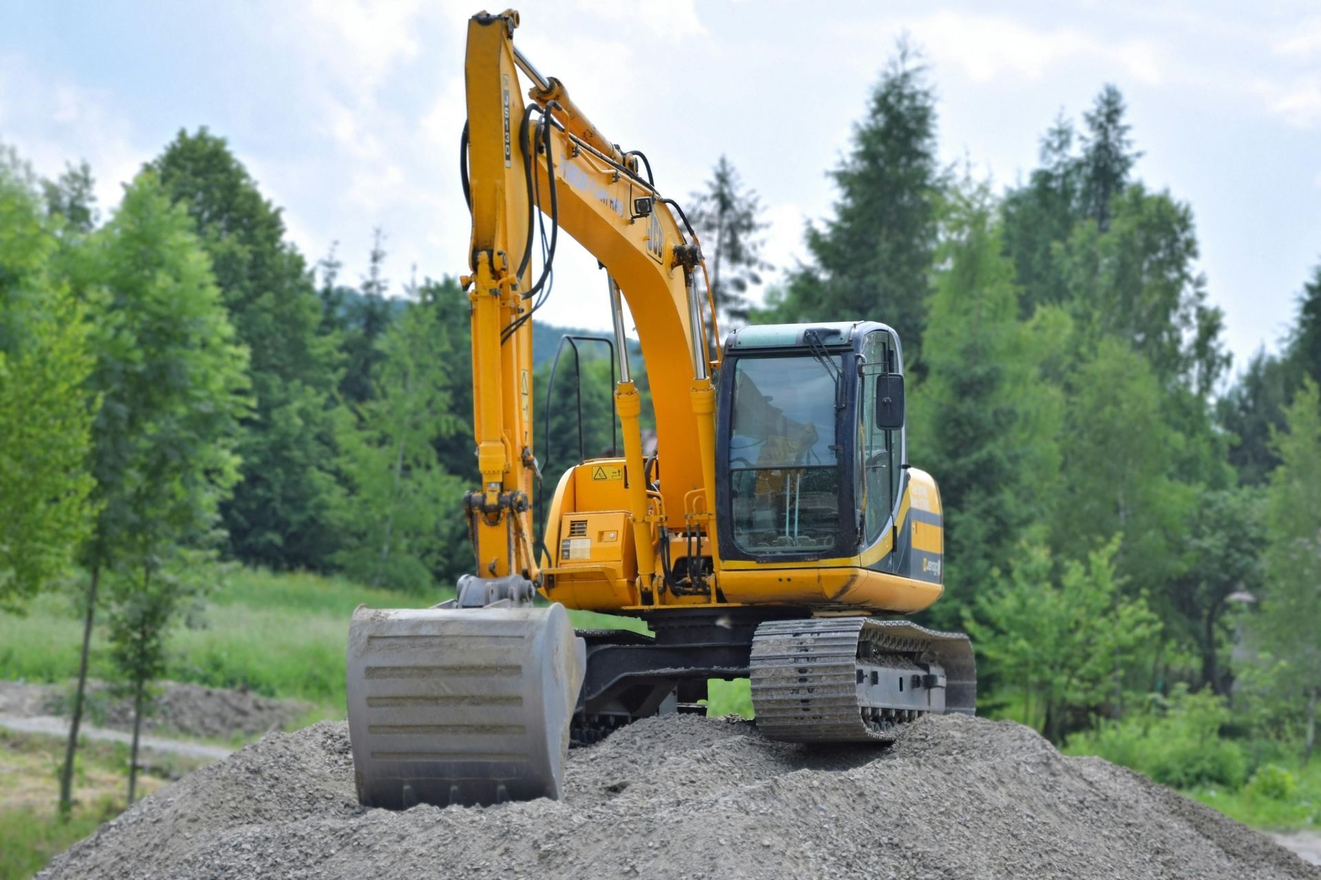 Yellow excavator on a pile of gravel, with green trees in the background.