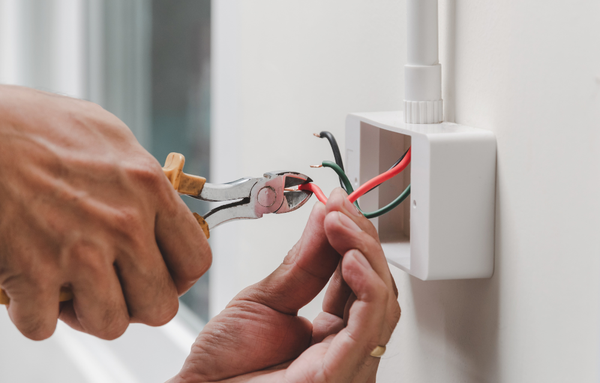 Persona cortando con alicates un cable eléctrico rojo en una caja de conexiones blanca. También se ve un cable verde.