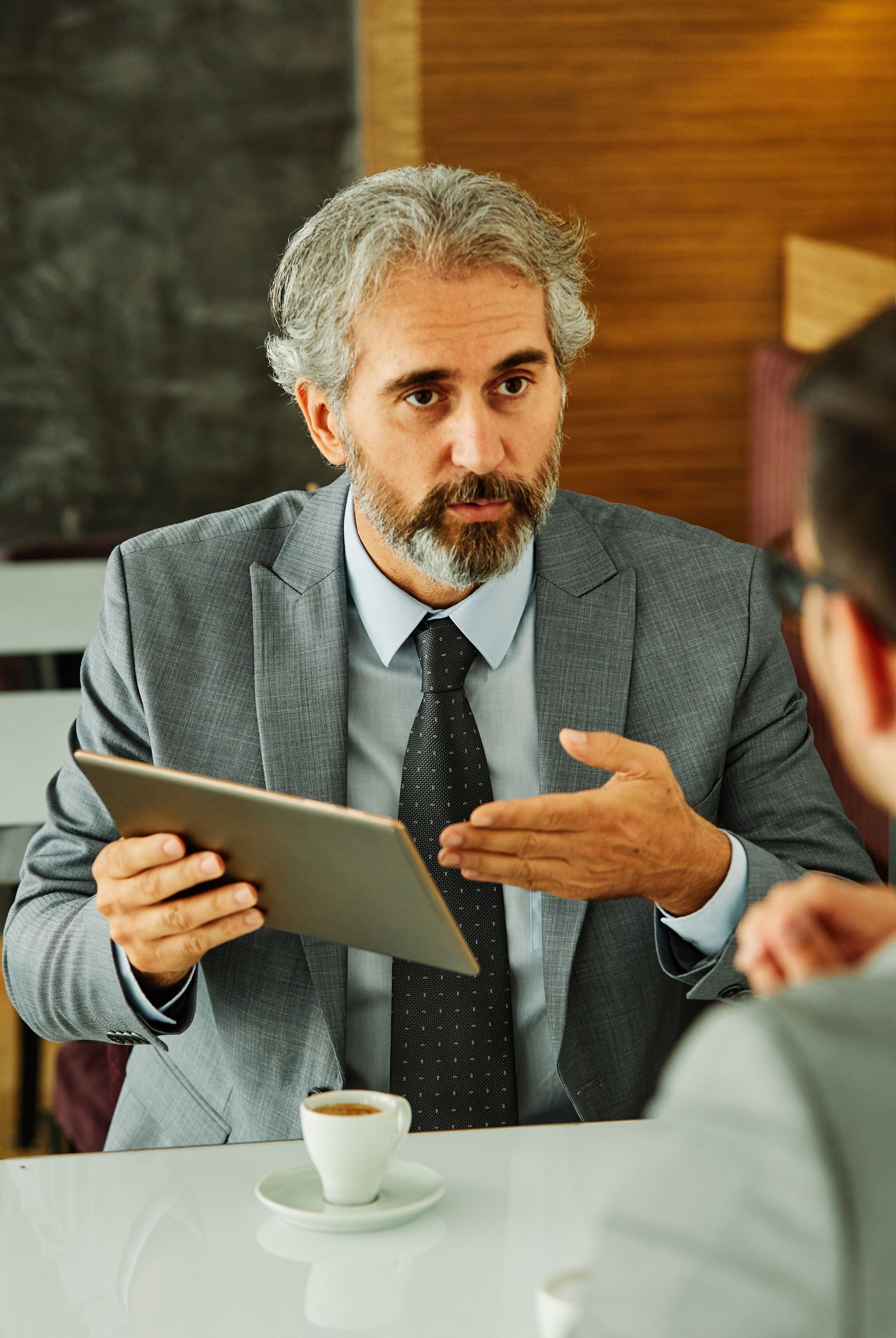 Man in suit gestures during a meeting, speaking to another person at a table.