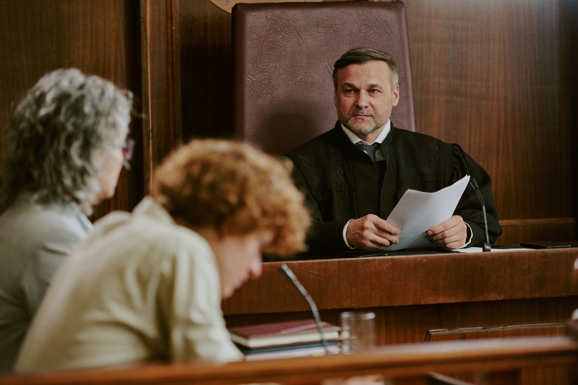 Judge in black robe, holding papers, presiding in a courtroom, with two women seated at a table.