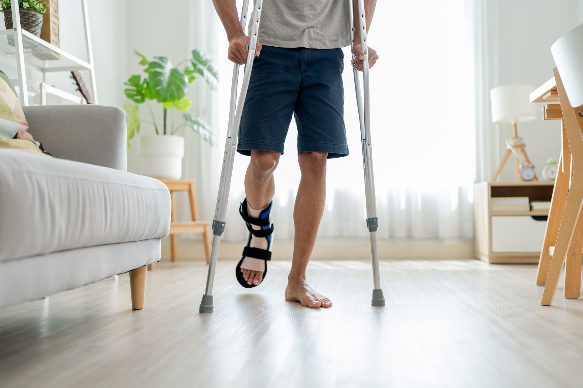 Man using crutches, leg in brace, walking indoors, light wood floors, near sofa and desk