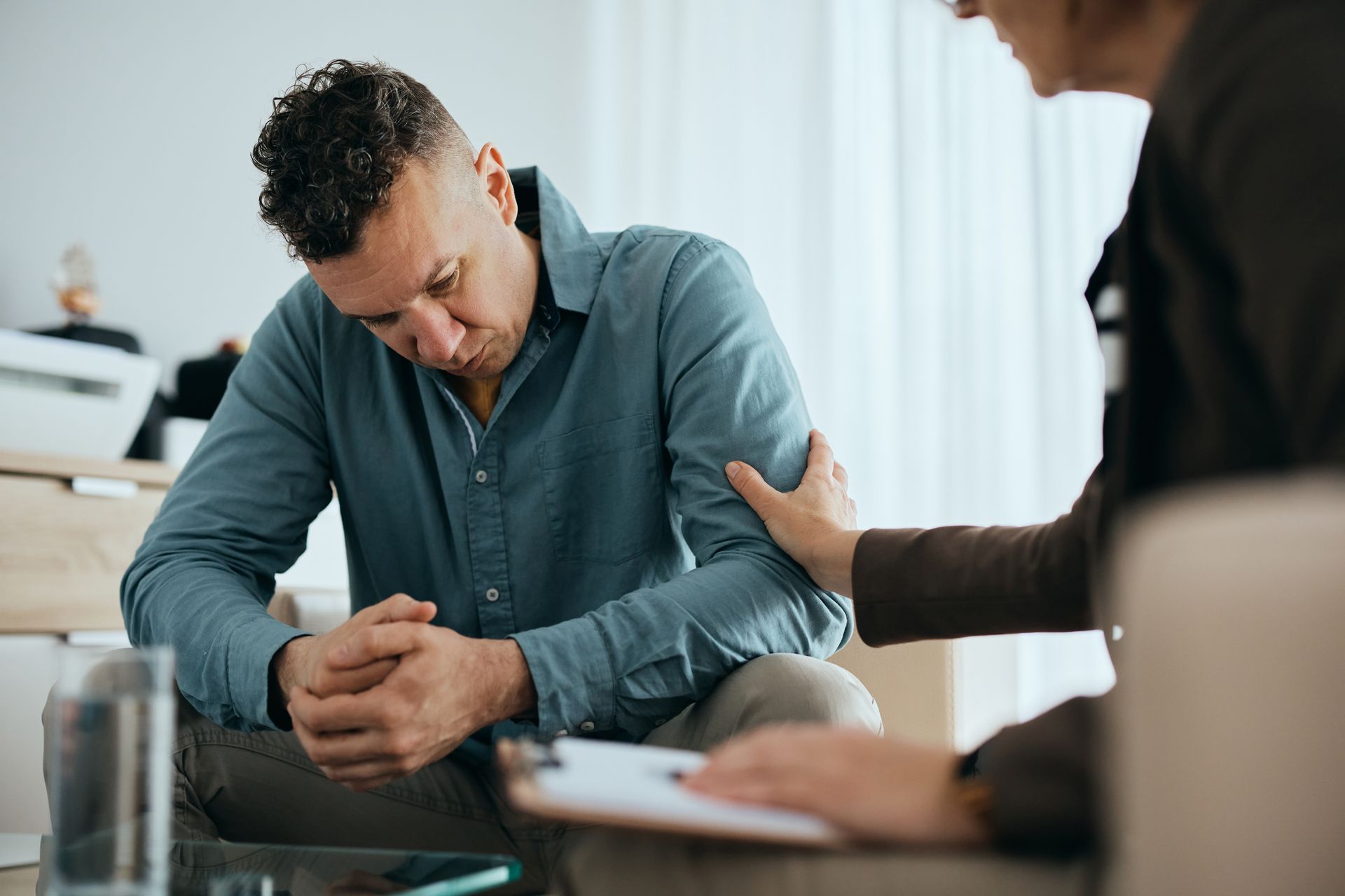 Man in therapy session, head down, being comforted by a person's hand on his shoulder; room setting.