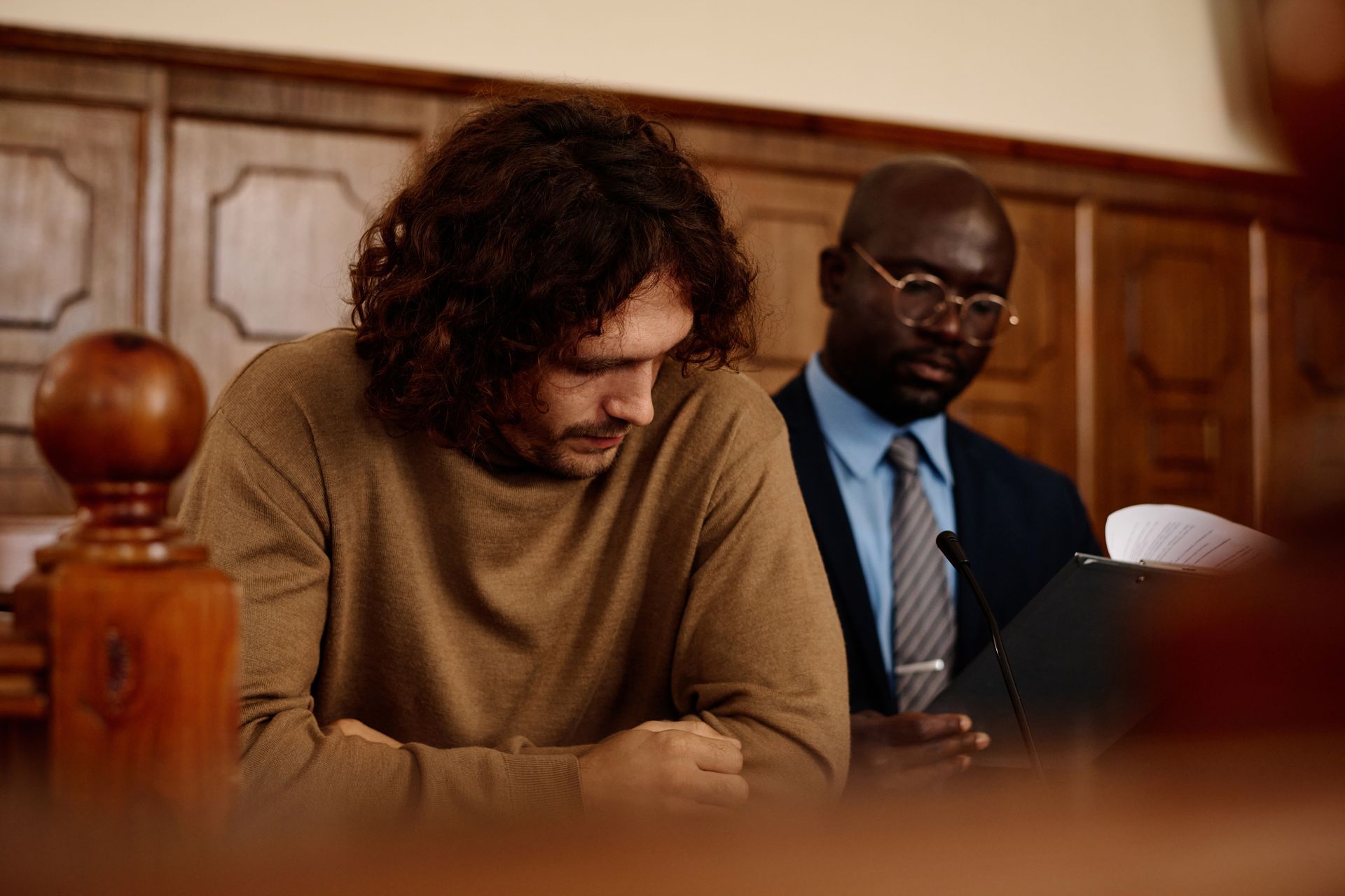Man in court with lawyer, head bowed, dark wood paneling, courtroom setting.