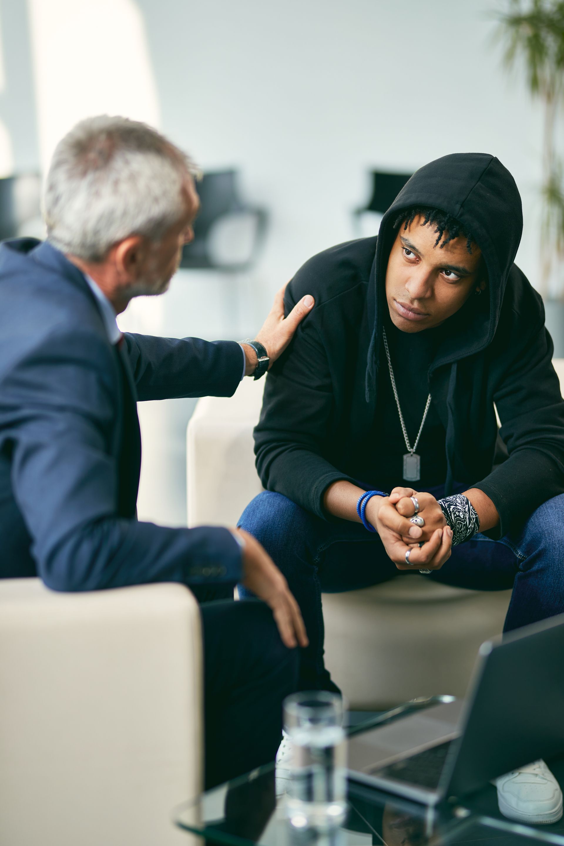 Man in suit comforts person in hoodie, both seated.  Modern office, laptop, glass of water on table.