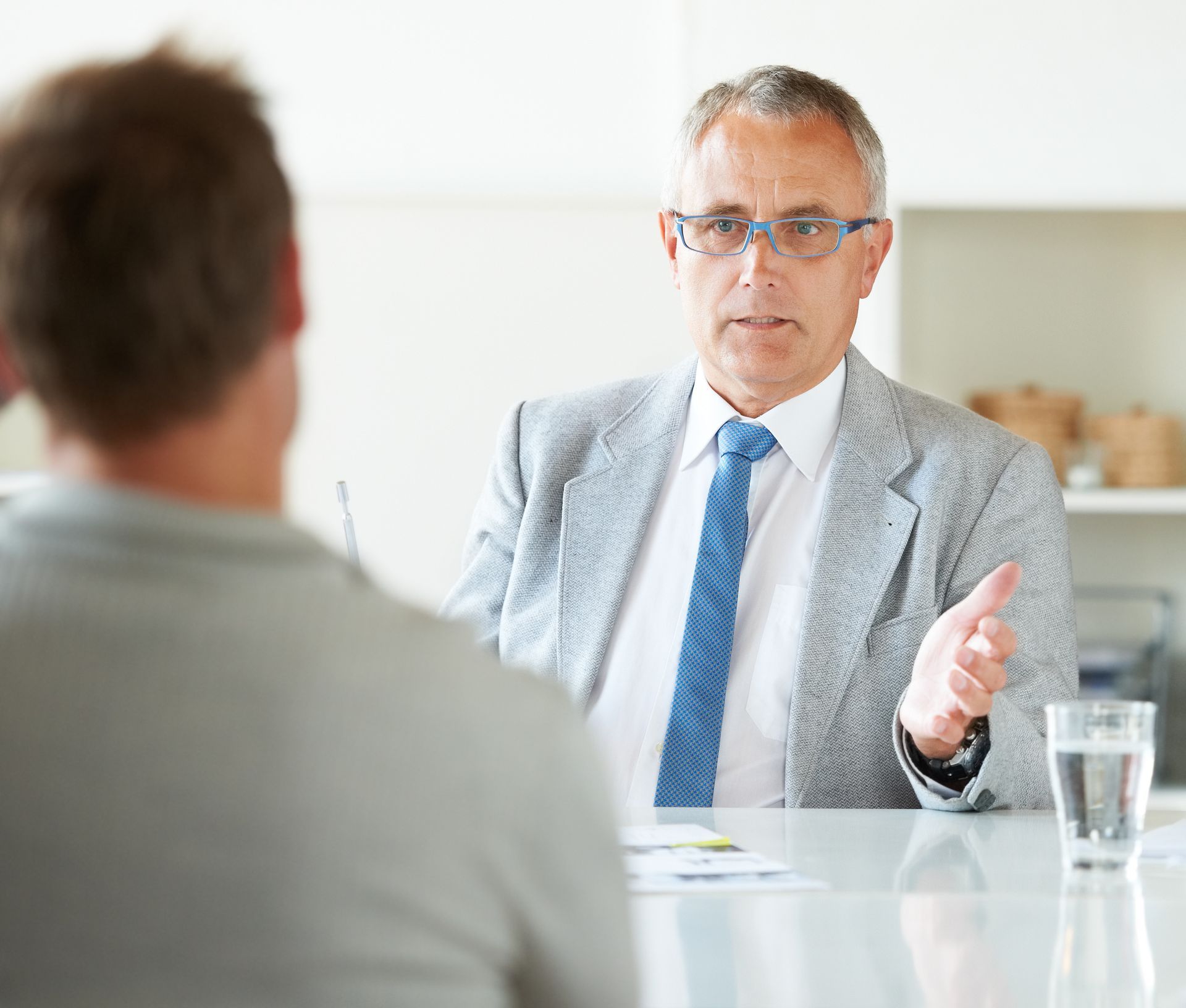 Man in suit gestures during a meeting, speaking to another person at a table.
