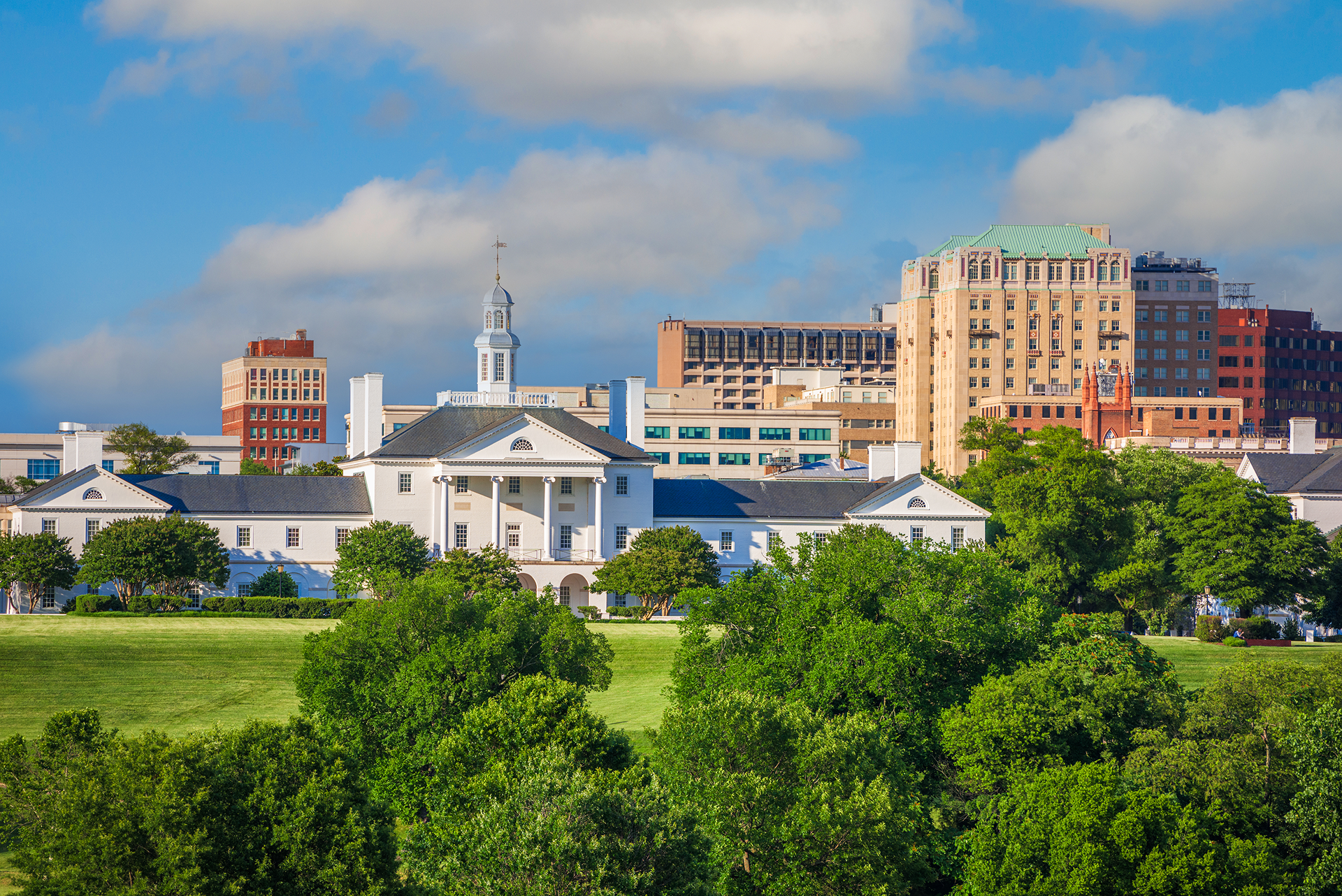 Skyline of Richmond, Virginia with state capitol building in foreground. Tall buildings in the background. Sunny day, trees.
