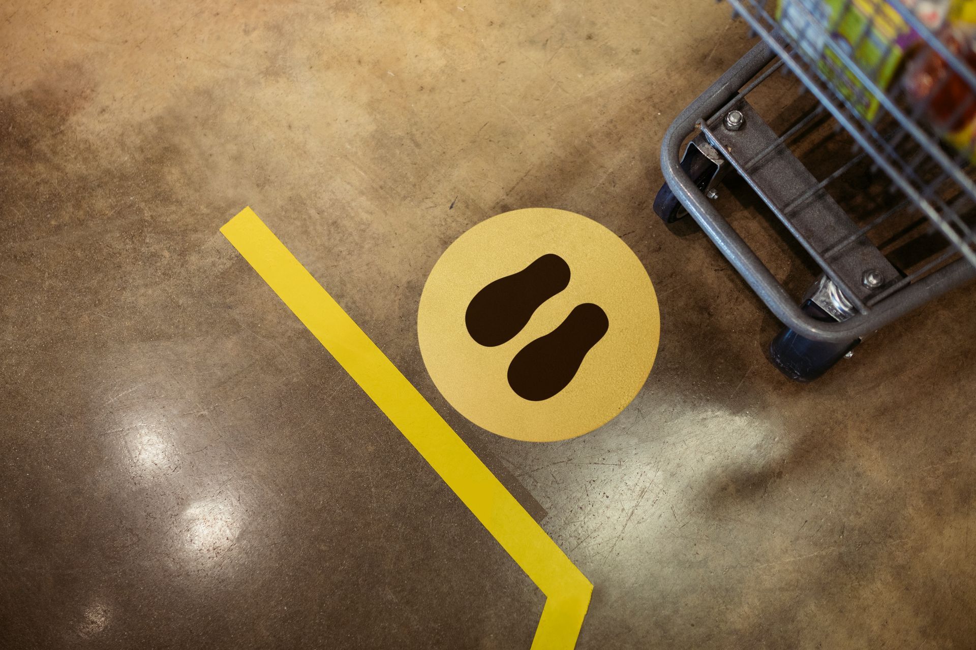 Yellow floor markings: footprints in a circle, and a line, near a shopping cart on a speckled floor.