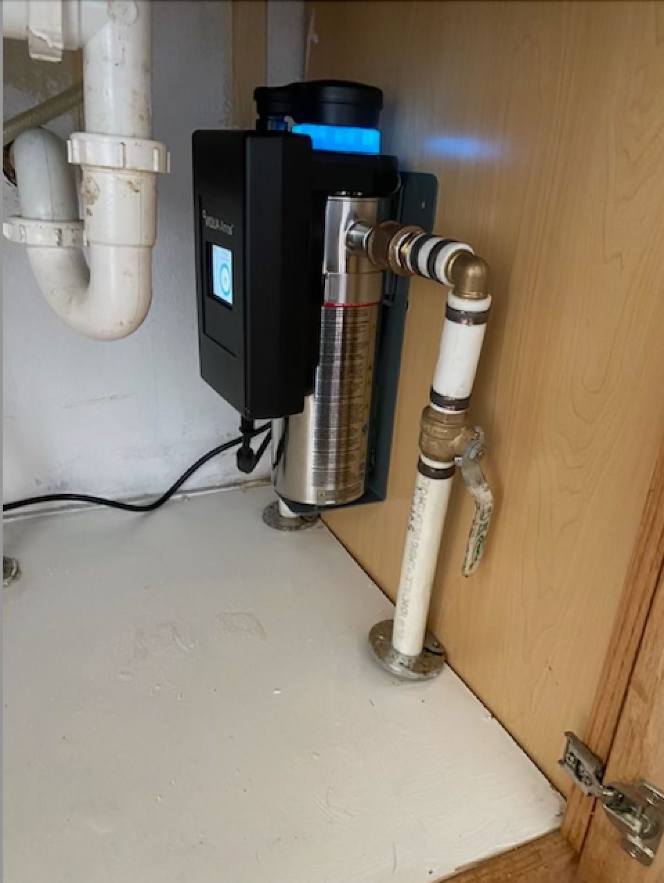 Under-sink water filtration unit with black housing, blue light, and connected pipes in a cabinet