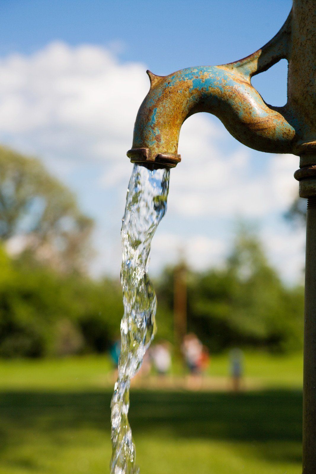 Water flowing from a rusty outdoor fountain spout into a park, with blurred trees and sky behind it