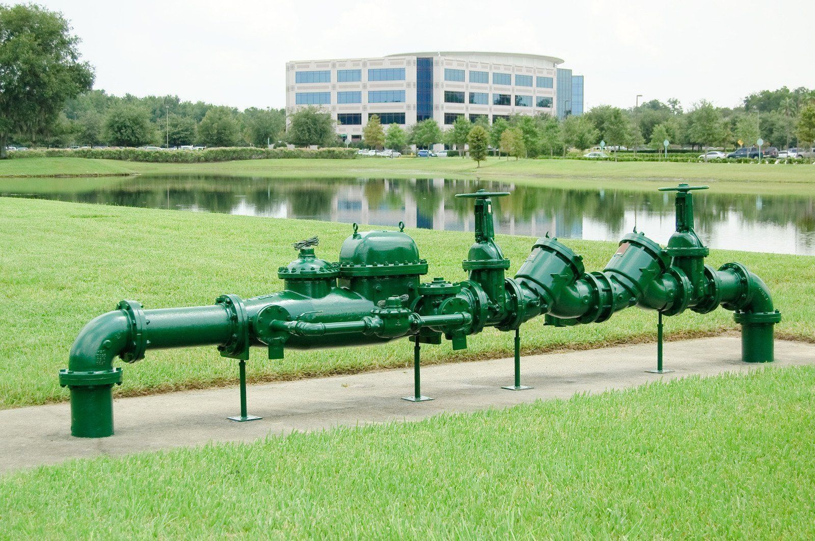 Green industrial pipeline and valves beside a pond in front of a modern office building