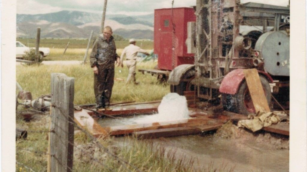 Person stands beside rustic machinery in a grassy field with mountains in the background.