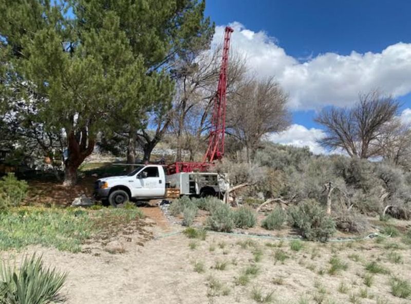 White truck and red drilling rig in a desert clearing with trees and brush under a blue sky