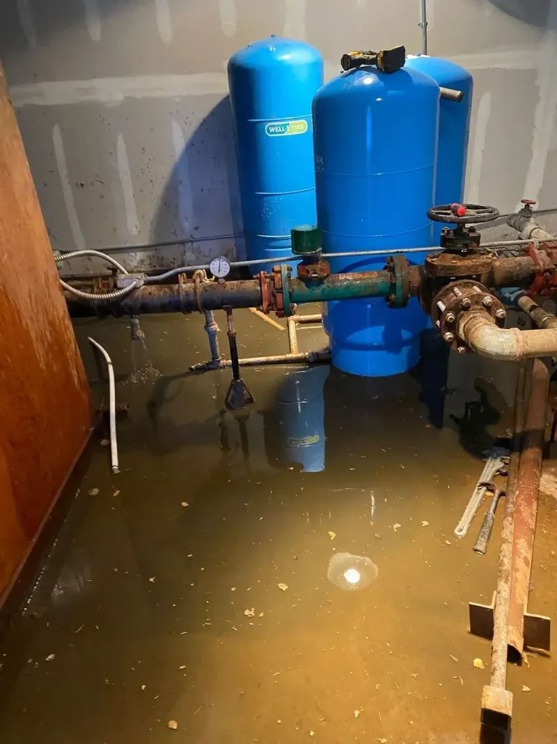 Flooded basement with blue water tanks, pipes, and standing water covering the floor