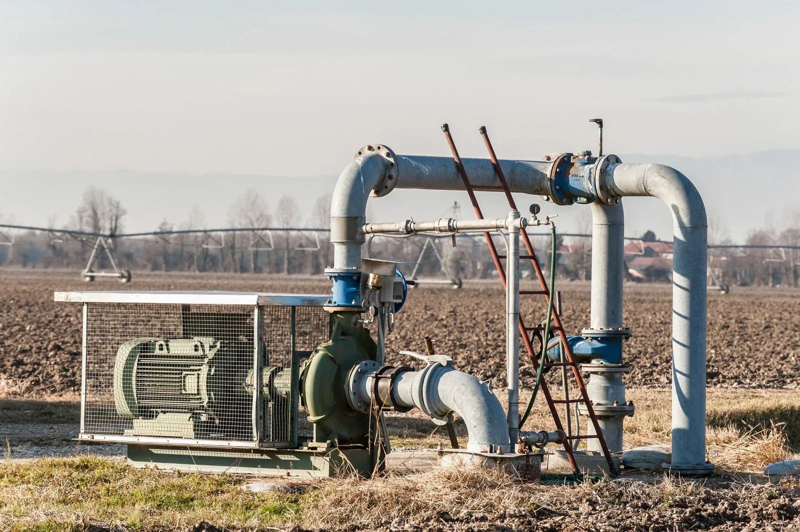Industrial pump and piping setup in a dry field, with blue and white pipes and a ladder.