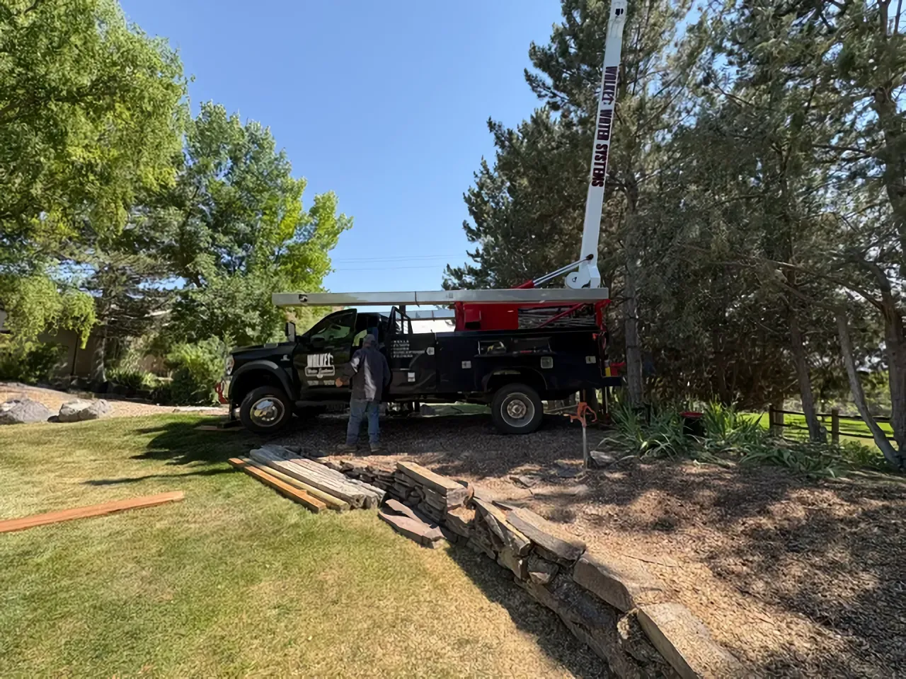Black utility truck with boom lift parked on a lawn near trees and a stone retaining wall