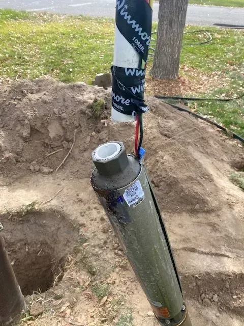 Broken utility pole base in a dirt hole, with exposed wires and plastic wrapping on a grassy roadside.