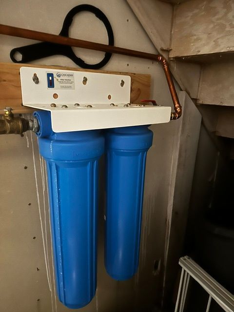 Blue whole-house water filter unit mounted under a sink, with two canisters and piping.