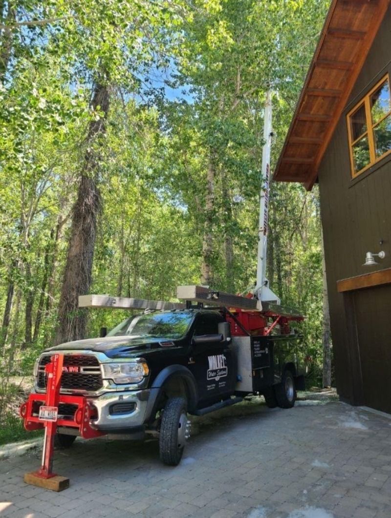 Black work truck with red equipment parked beside a brown building in a wooded driveway