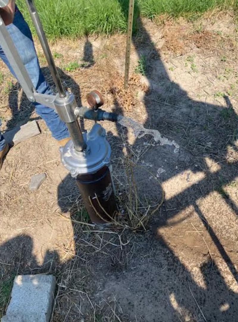 Hand pump beside a black water well in a grassy outdoor area, with shadows on the ground