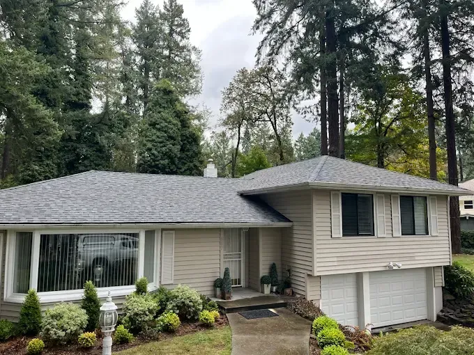 Beige house with gray roof, surrounded by green trees and landscaping, overcast sky.