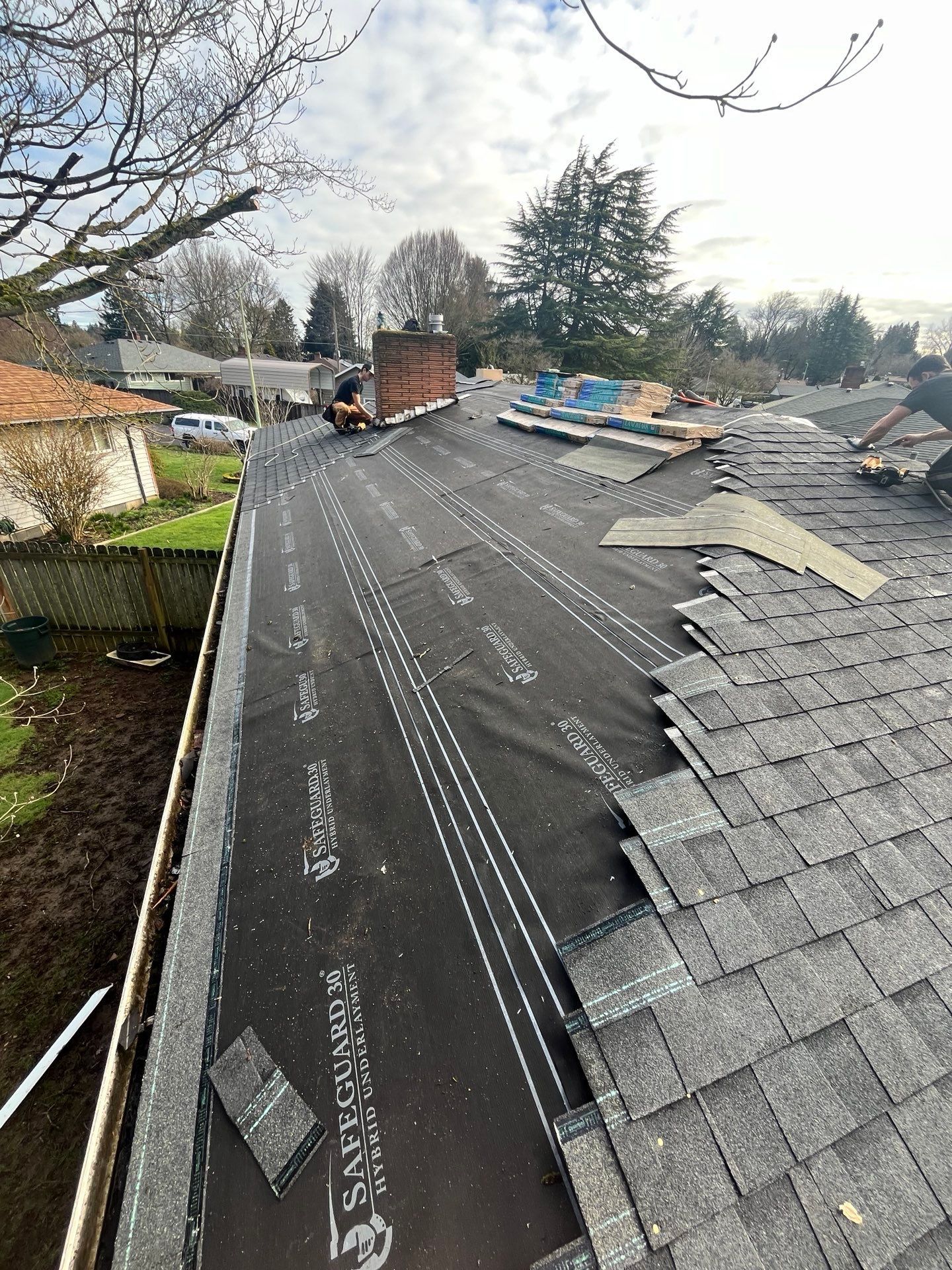 Roof partially covered with black underlayment, shingles, and workers near the chimney.