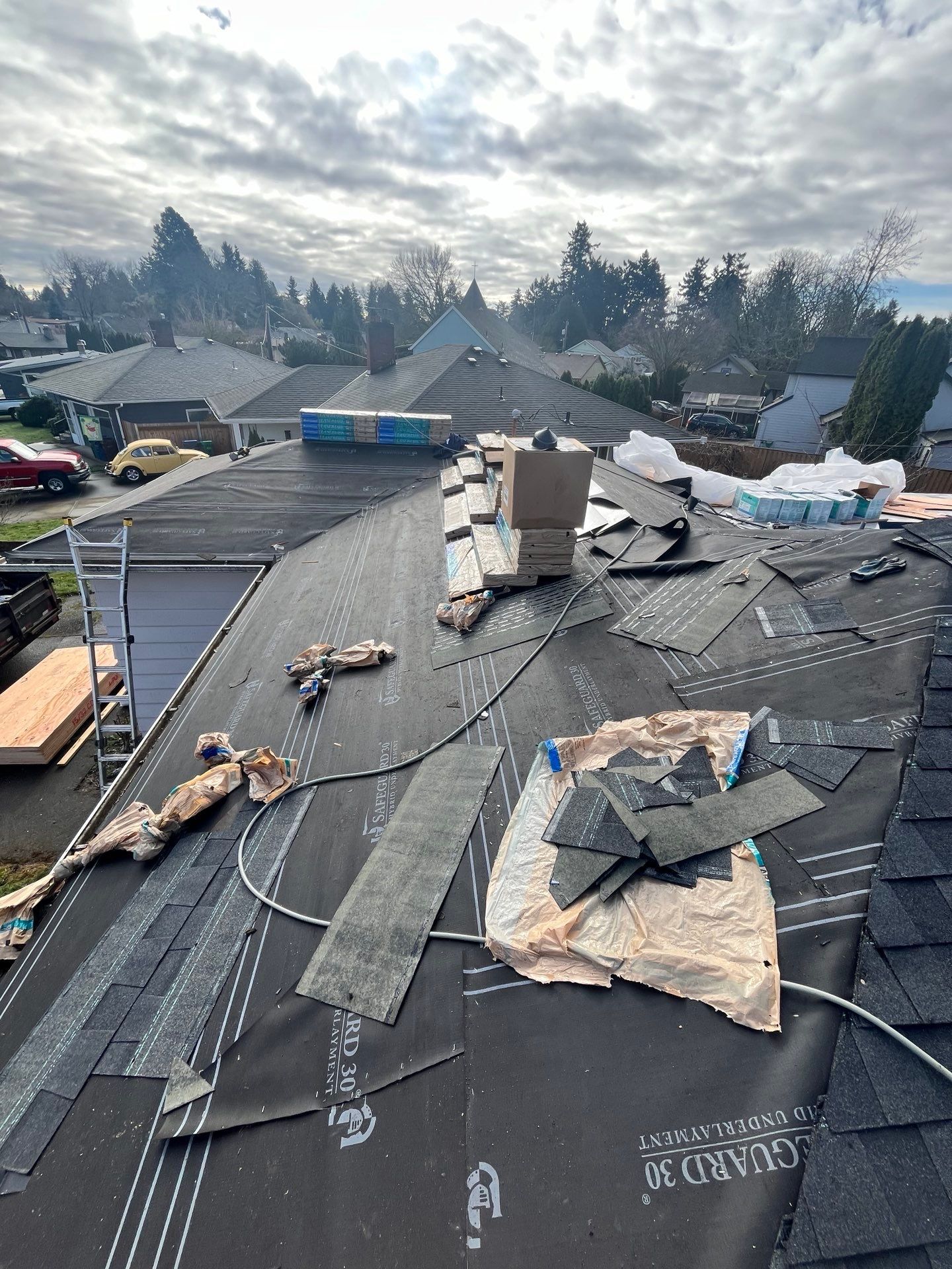 Rooftop with partially removed shingles, underlayment exposed. Chimney and surrounding homes visible. Overcast sky.