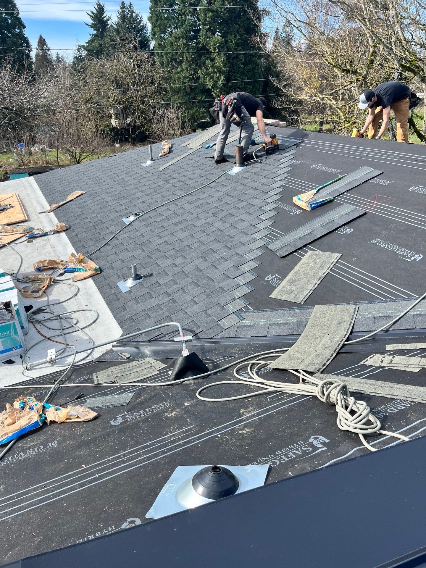 Two workers install shingles on a roof. One cuts shingles, the other gathers debris. Sunny day, trees in background.