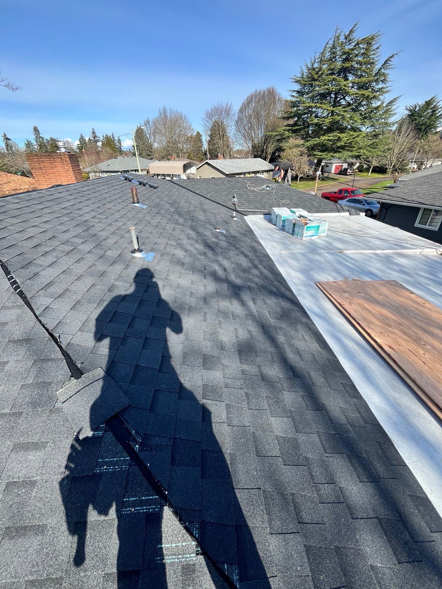 Man's shadow on a newly shingled roof under a clear, blue sky. Construction supplies are visible.