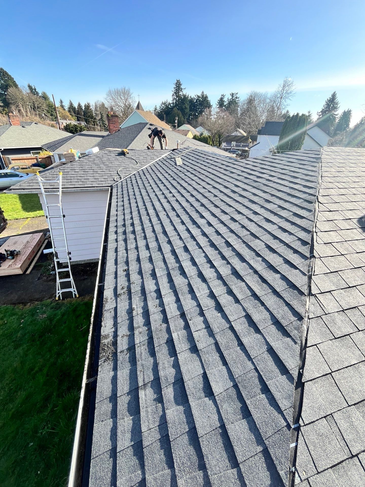 View of a dark gray shingle roof on a house, with a ladder leaning against it on a sunny day.