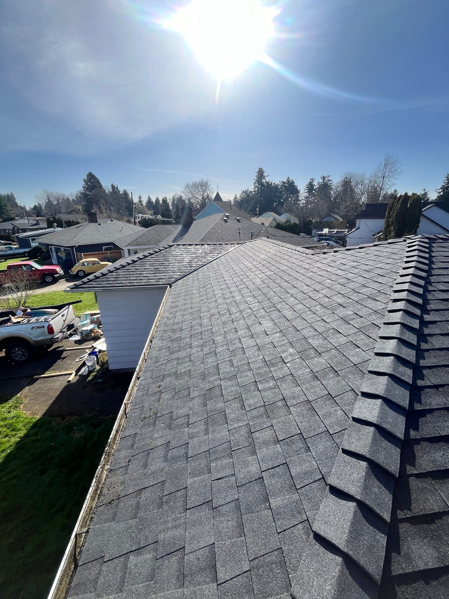Rooftop view of gray shingle roof under a bright sunny sky with surrounding houses and trees.
