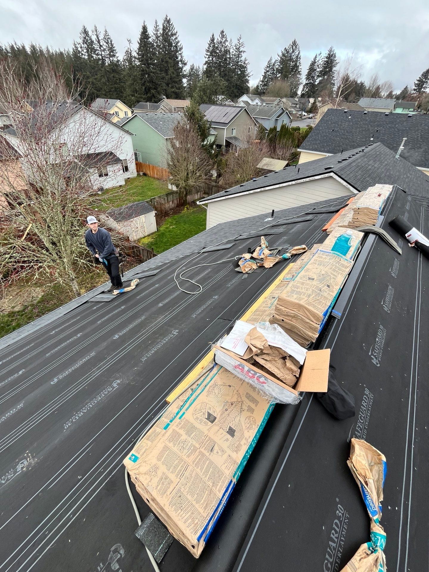 Man on a roof installing shingles, with bundles of roofing materials. Cloudy day, houses in background.