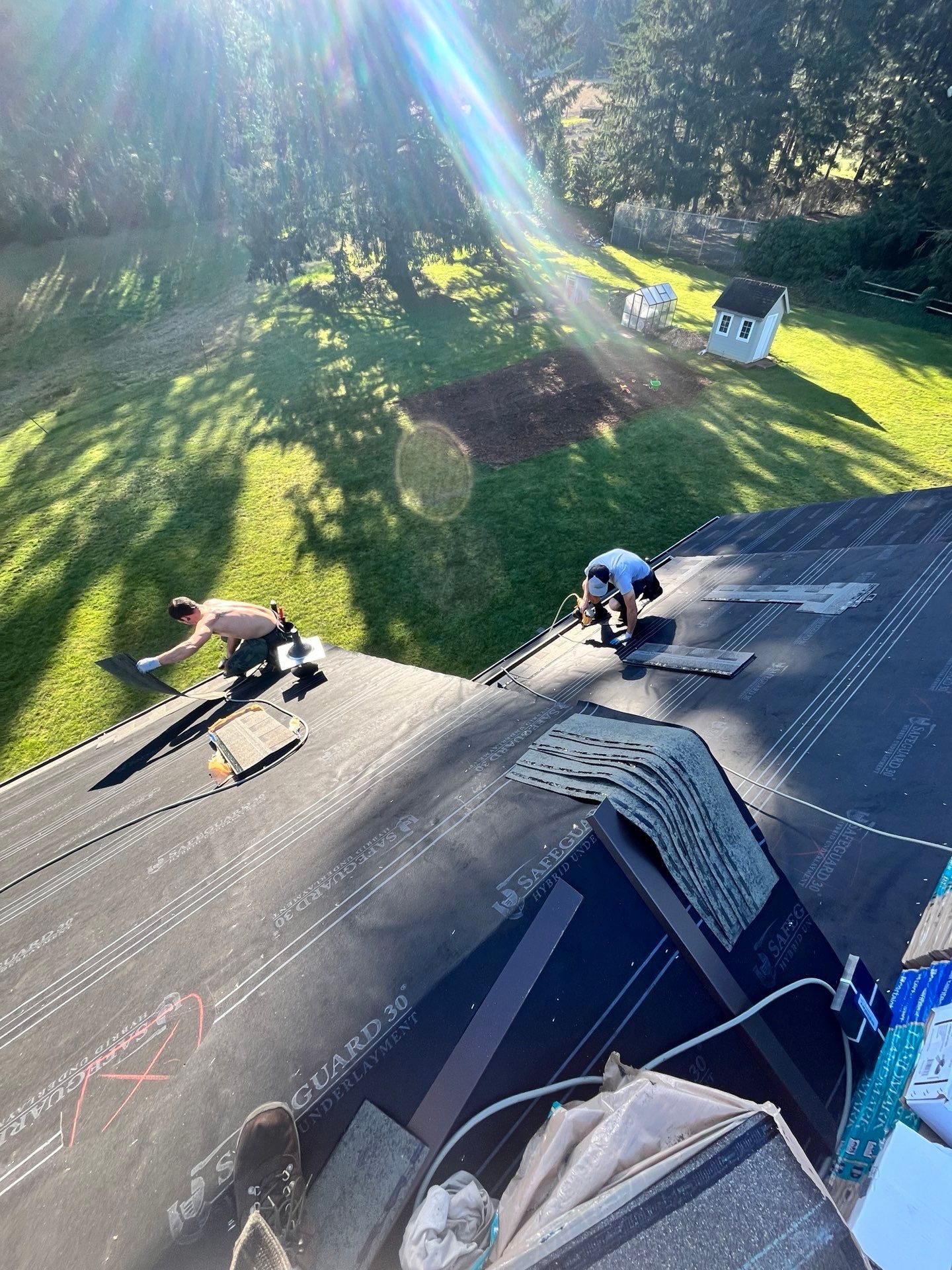 Two workers install roofing material on a sunlit roof; green lawn and trees in the background.