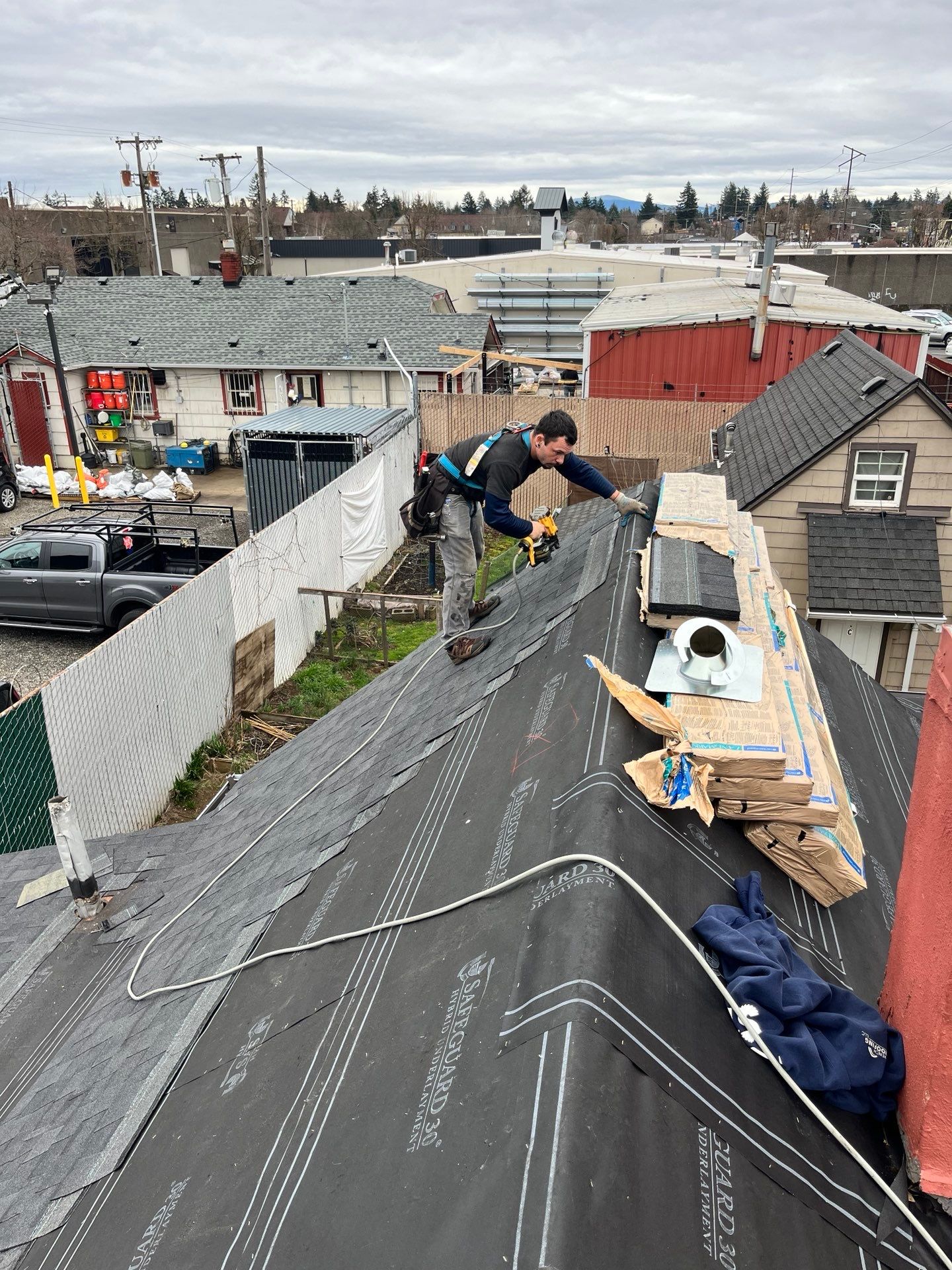 Roofer working on a dark roof, holding a tool, surrounded by materials, in a residential area.
