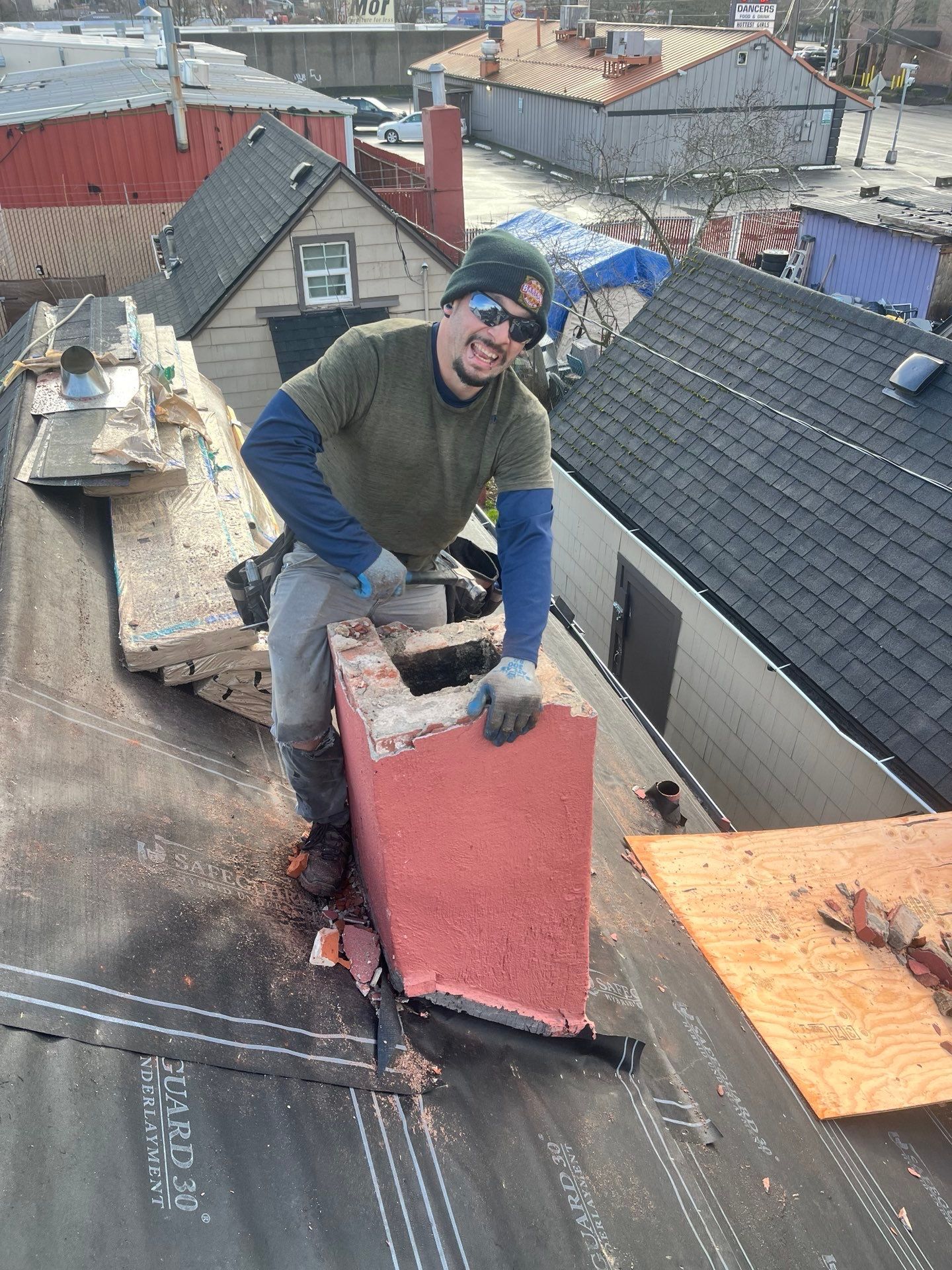 Person on a rooftop smiles, holding a red chimney piece. Construction site, bright day.