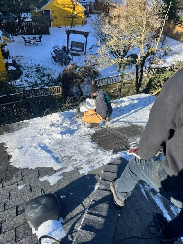 Workers clearing snow from a rooftop. Snow-covered yard in the background. Sunny day.