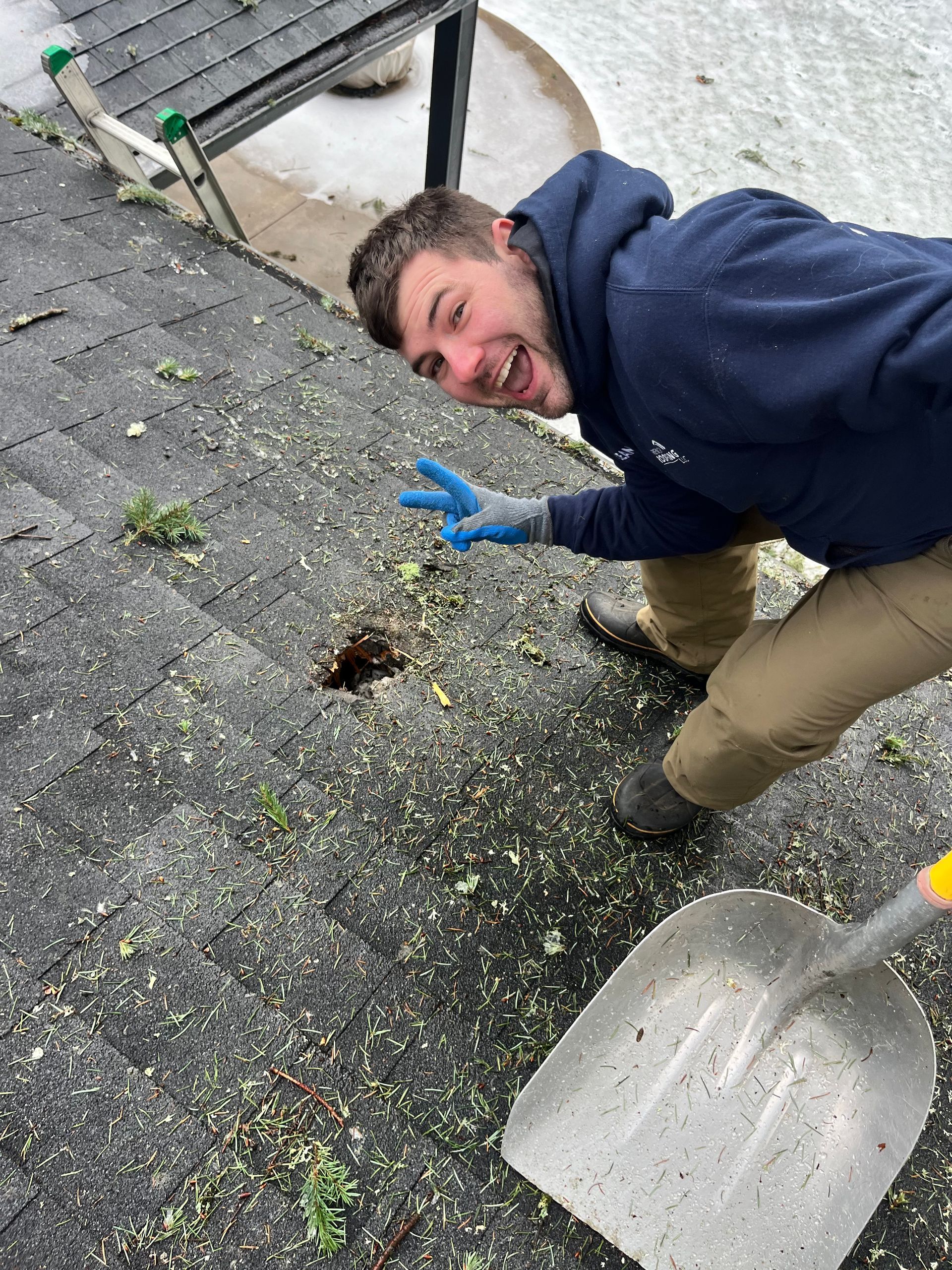 Man on rooftop with shovel, smiling, pointing with two fingers, roof damaged with debris and snow.