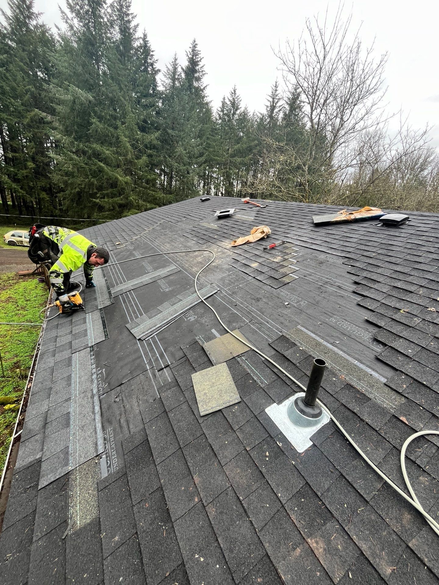 Two workers on a roof, replacing shingles. One cuts with a saw. Grey and black shingles, green grass, and trees.