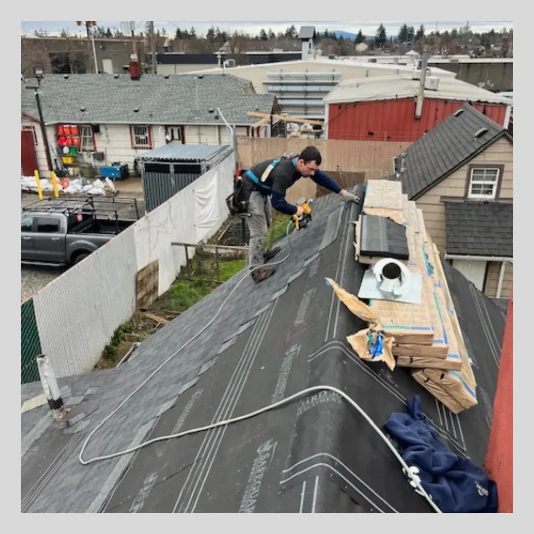 Roofer working on a dark roof, holding a power drill. He is wearing a safety harness. Other buildings are in the background.