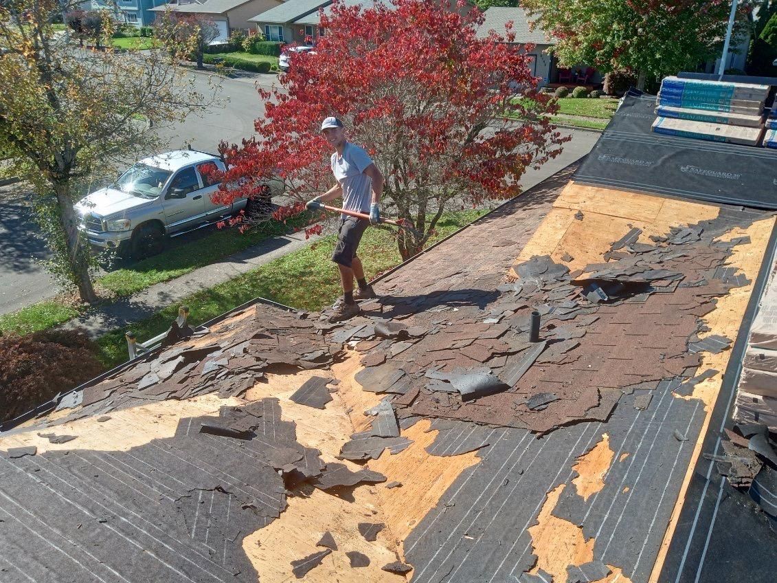 Person removing shingles from a roof. Brown and yellow debris on the roof, with a car and trees in the background.
