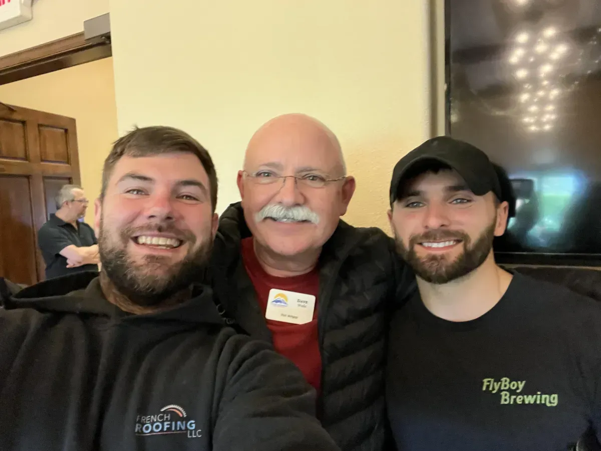 Three men smiling, posing for a photo indoors. Two on either side of a man with a mustache.