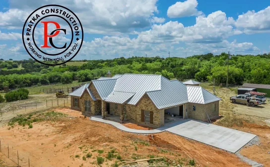 A partially built stone house with a concrete driveway, set against a landscape with trees, under a cloudy sky.