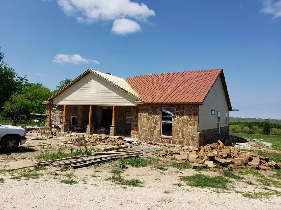 House under construction with stone facade, red roof, beige siding, and blue sky.