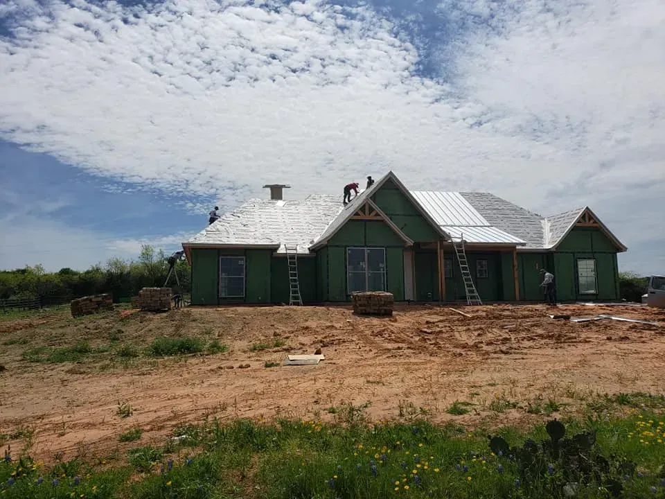 Construction of a house with a metal roof, workers on roof, green siding, on a dirt lot under a cloudy sky.