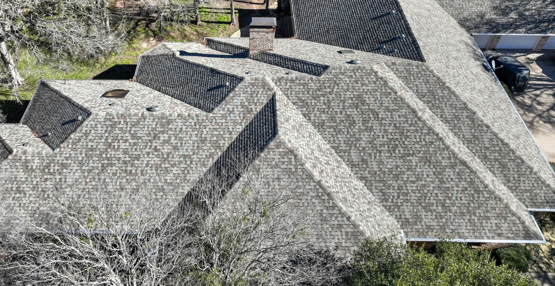 Overhead view of a gray shingle roof with multiple intersecting planes; trees in the foreground.