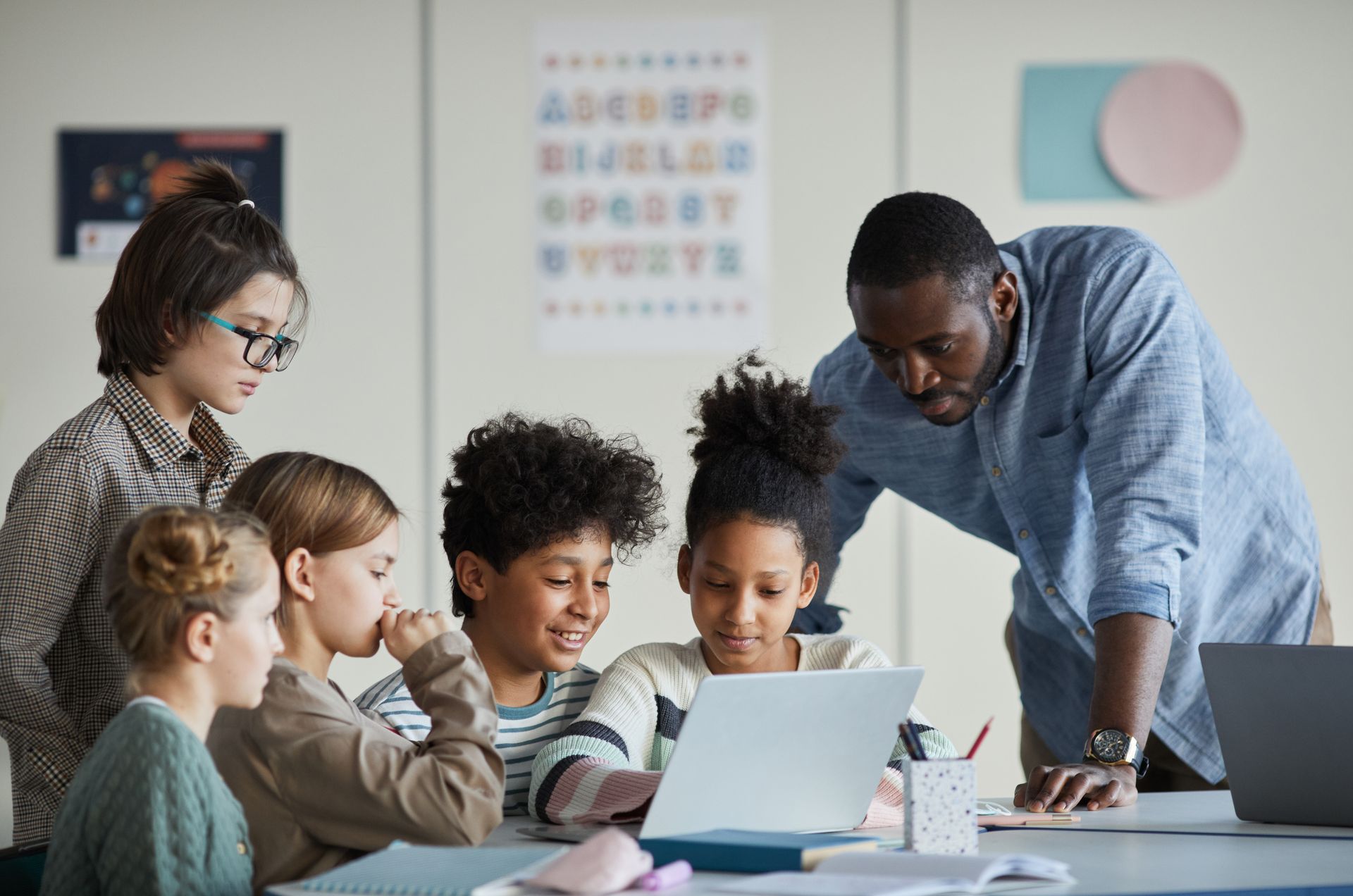 A teacher leans over a table, guiding a diverse group of engaged children gathered around a laptop in a classroom.