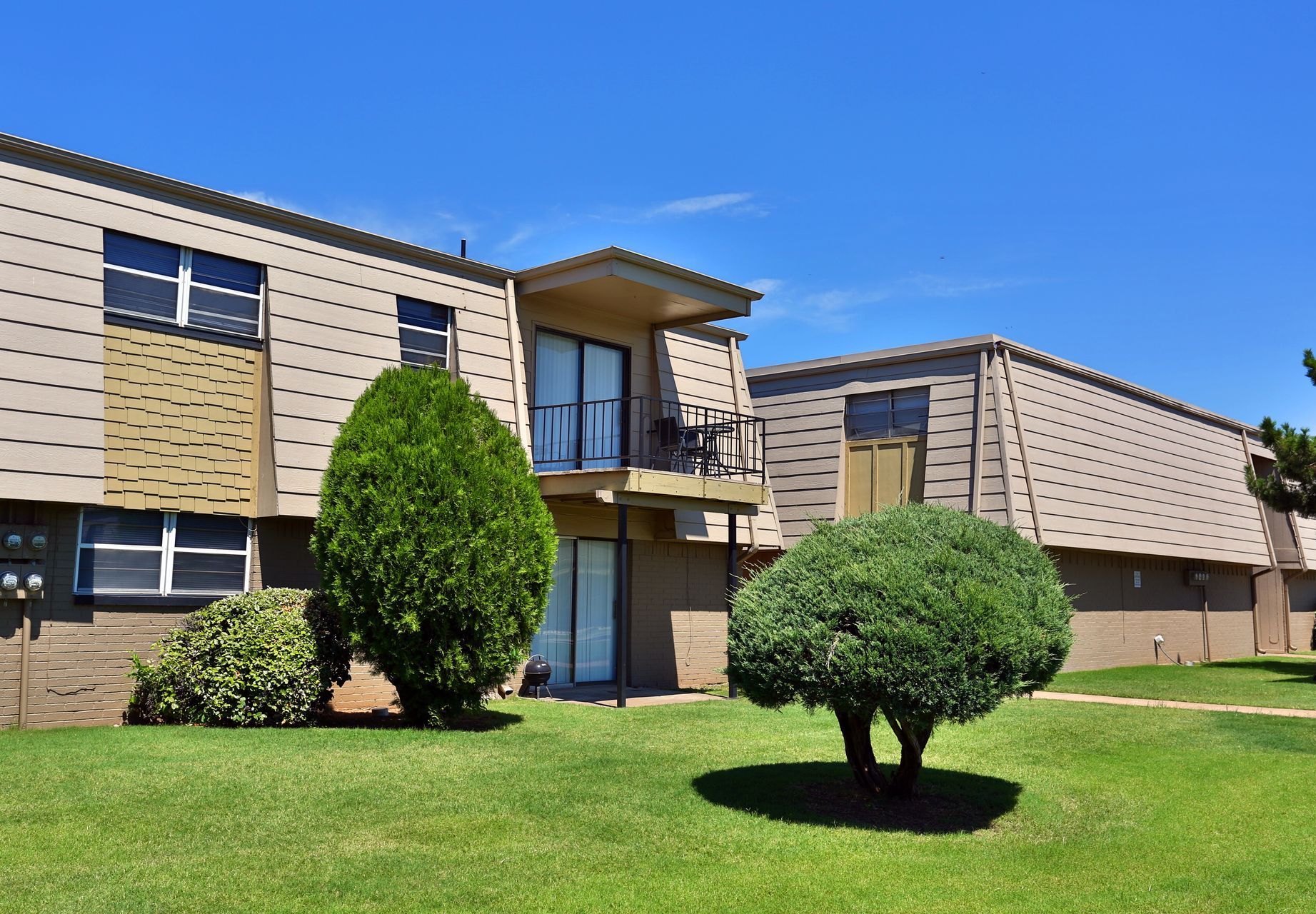 Two-story apartment buildings with beige siding, balcony, green lawn, and trees under a blue sky.