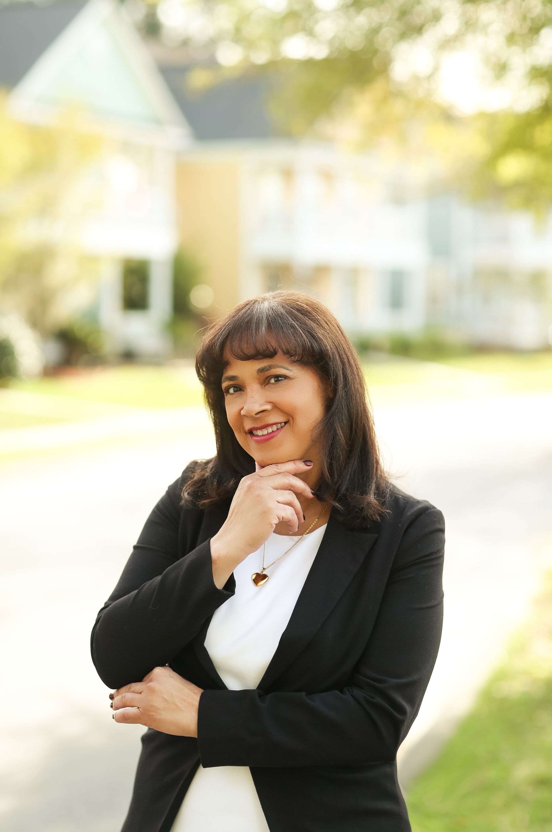 A woman in a black jacket and white shirt is standing in front of a house.