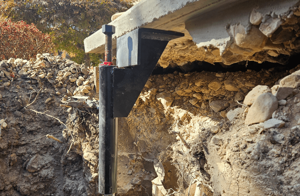 A foundation pier supports a concrete structure in a trench. The pier is black metal, with dirt and rocks surrounding.